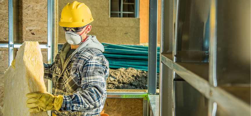 Construction worker in safety helmet and respirator mask fitting yellow batt commercial insulation into steel stud framing.